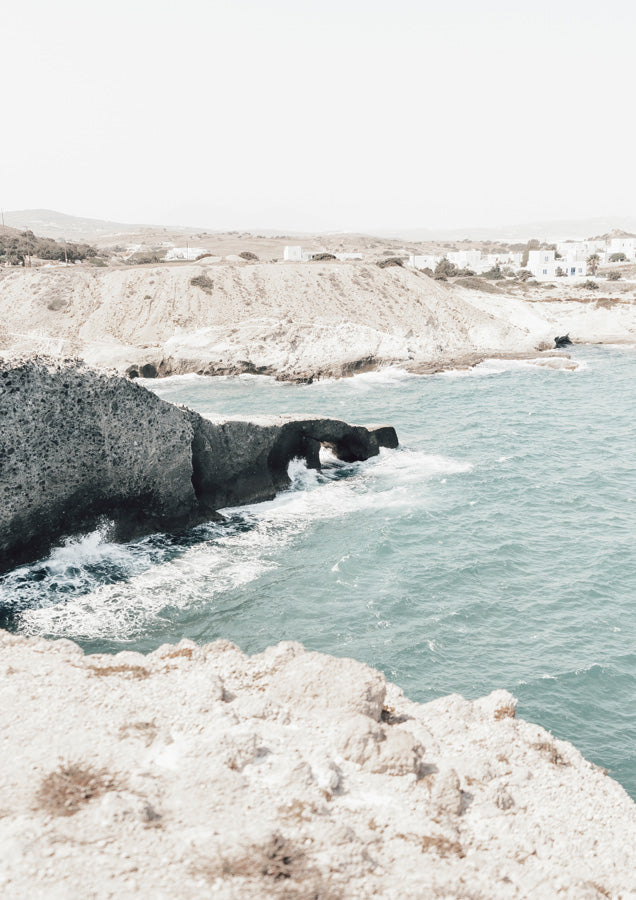 Dark rocky coastline with arch formation and turquoise sea near a whitewashed village on Milos