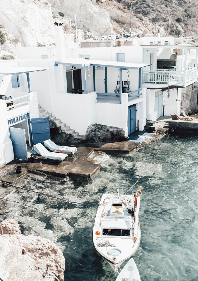 Whitewashed Greek village buildings with blue shutters above turquoise water and a moored boat