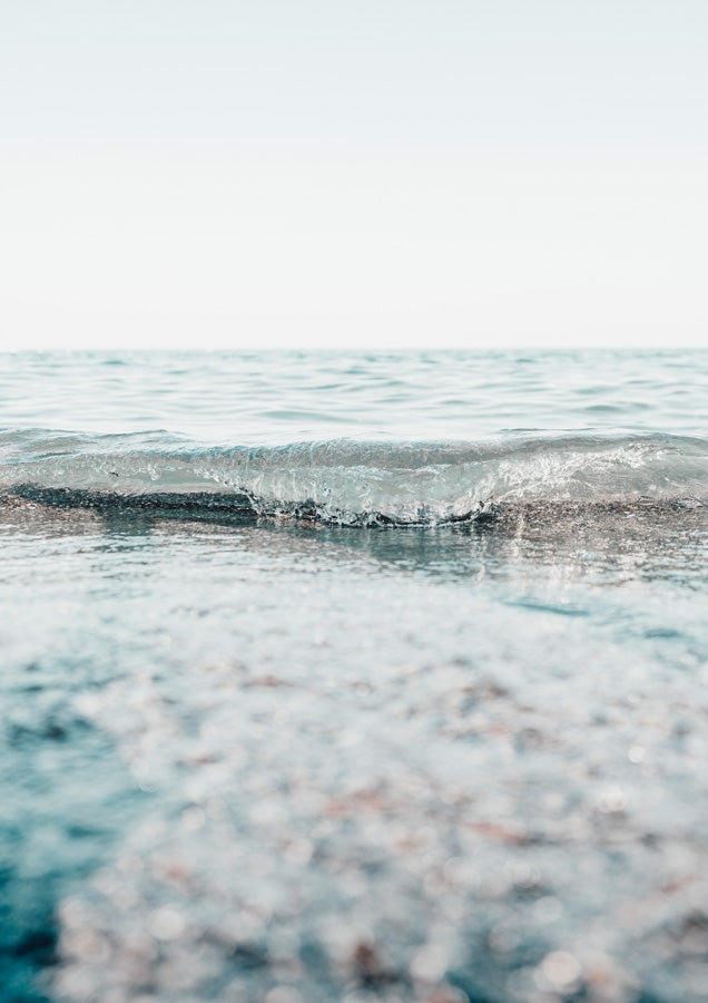 Close-up photograph of a breaking ocean wave with soft-focus water and pebbles in pale blue tones